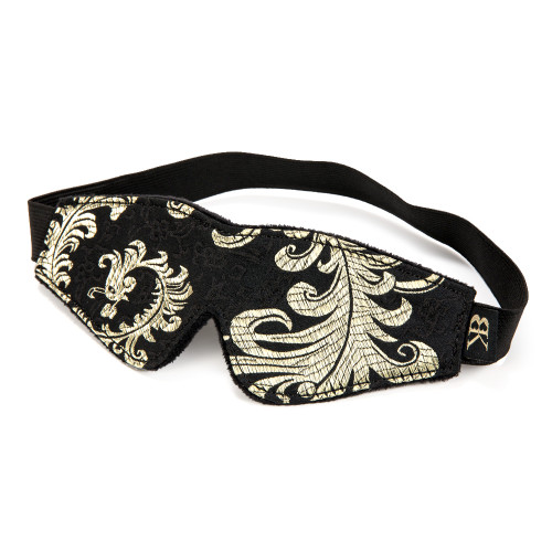 An angled view of the Dark Desire eye mask from our Dark Desires Collection. The mask has a black and gold filigree pattern and an elastic strap to fit around the head. It sits on a white background