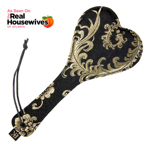 An image of the Heart's Desire heart-shaped spanking paddle from the Dark Desires collection lying on a white background. The paddle is covered in black and gold filigree fabric and has a cord.