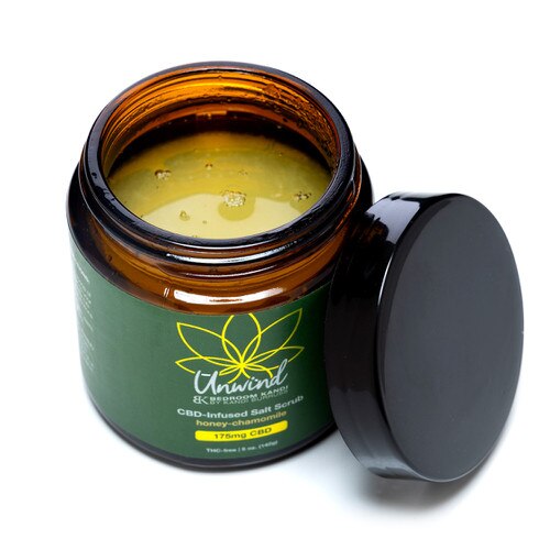 A view of an open jar of Unwind Salt Scrub with the black lid beside it on a white background